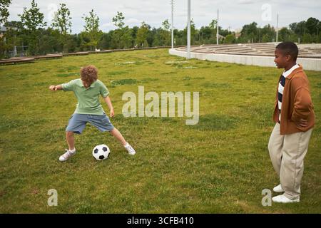 Kaukasischer Junge, der Fußball auf dem Gras dribbelt, während Black Boy in der Nähe beobachtet wird, während beide Kinder tagsüber im Park sportlich aktiv sind Stockfoto