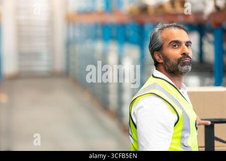 Lagerarbeiter mittleren Alters schiebt den Transportwagen mit Kiste und trägt eine Sicherheitsweste im Gang, Kopierraum Stockfoto