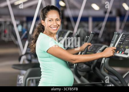 Schwangere Frau, die im Fitnessstudio mit Konsole und Kardiogeräten auf einem Crosstrainer steht Stockfoto