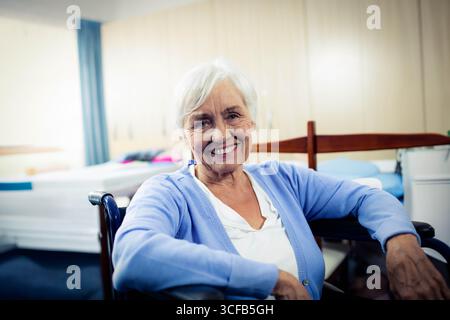 Seniorin sitzt im Rollstuhl im Pflegeraum neben Bett mit Geländer und blauem Vorhang Stockfoto
