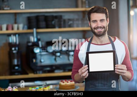 Bärtiger Mann Barista, der ein Tablet hält, das im Café mit Gebäck und Espressomaschine steht Stockfoto