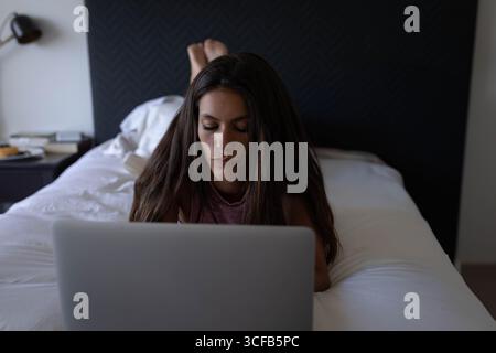 Frau, die auf dem Bauch auf dem Bett lag und sich auf einen Laptop konzentrierte, in einem modernen Schlafzimmer mit Nachttisch Stockfoto