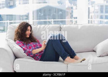 Frau, die sich auf einem hellgrauen Sofa zurücklehnt und ein Tablet in einem modernen Wohnzimmer mit Stadtblick benutzt Stockfoto