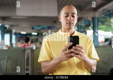 Reifer kahlköpfiger hispanischer Mann, der an einem Busbahnsteig im Freien sitzt und auf den Transport wartet, während er sein Smartphone durchsucht. Reisen, Pendeln und Stockfoto