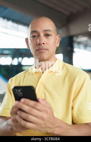 Reifer kahlköpfiger hispanischer Mann, der an einem Busbahnsteig im Freien sitzt und auf den Transport wartet, während er sein Smartphone durchsucht. Reisen, Pendeln und Stockfoto