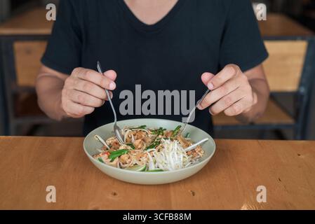 Porträt eines hispanischen Touristen mittleren Alters, der traditionelle Thai Pad Thai Nudeln in einem Restaurant im Freien genießt und dabei lächelt, während dini Stockfoto