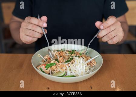 Porträt eines hispanischen Touristen mittleren Alters, der traditionelle Thai Pad Thai Nudeln in einem Restaurant im Freien genießt und dabei lächelt, während dini Stockfoto