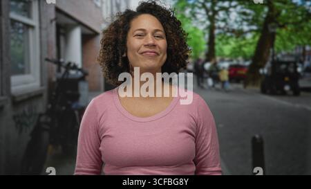 Frau in rosa Pullover gibt auf der belebten Stadtstraße, die von Bäumen und geparkten Fahrrädern gesäumt ist, zwei Daumen hoch; Optimismus. Stockfoto