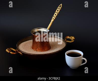 Kupfertürke in einem Tablett mit Sand und einer Tasse heißem frisch gebrühtem schwarzem Kaffee auf schwarzem Hintergrund. Nahaufnahme. Stockfoto