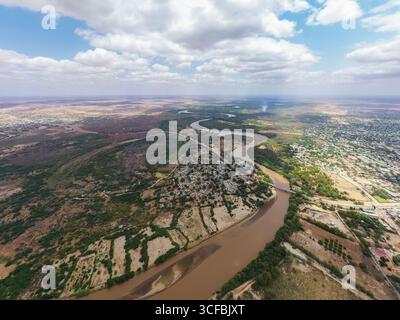 Aus der Vogelperspektive auf den braunen, gewundenen Tana River, der sich durch eine Landschaft mit Siedlungen und grüner Vegetation schlängelt, Garissa, Kenia. Stockfoto