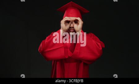 Junger chinesischer Mann in roter Graduierungsmütze und rotem Kleid macht binokulare Gesten isoliert auf schwarzem Hintergrund, die Neugier und Zukunftsvision symbolisieren. Stockfoto