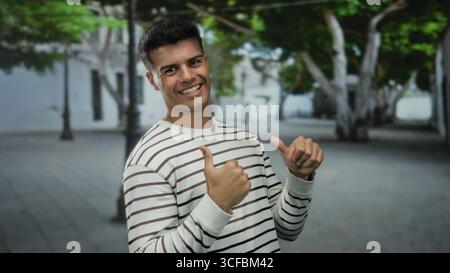 Ein junger Mann im Freien, der auf einer Stadtstraße mit Bäumen und Gebäuden im Hintergrund die Daumen hochlegt, ein gestreiftes Hemd trägt und selbstbewusst lächelt, verschwindet Stockfoto