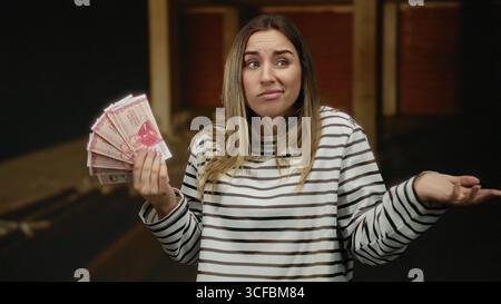 Frau mit isländischen Kronen-Banknoten auf einer Innenbaustelle mit einem verwirrten Ausdruck, der den unvollendeten Hintergrund kontrastiert. Stockfoto