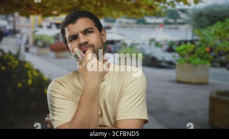 Junger hispanischer Mann mit beigefarbenem T-Shirt, der zerknitterte Riyal-Banknoten gegen das Kinn auf der belebten Straße hält; Nachsicht. Stockfoto