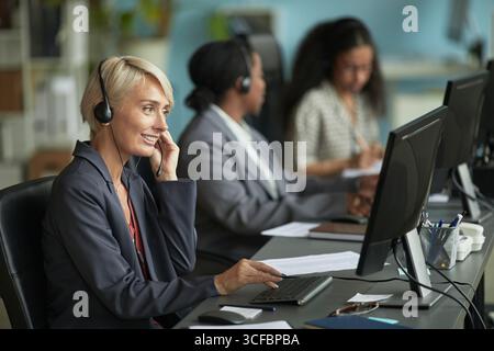 Kaukasische Frau mittleren Alters, die ein Headset trägt, in einem modernen Büro am Computer arbeitet, mit zwei Kollegen im Hintergrund mit Computern und Headsets am Schreibtisch sitzt Stockfoto