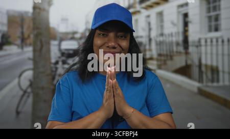 afroamerikanerin, die eine blaue Mütze mit zusammengedrückten Palmen trägt, lächelt selbstbewusst auf einem Bürgersteig in der Stadt; Dankbarkeit. Stockfoto