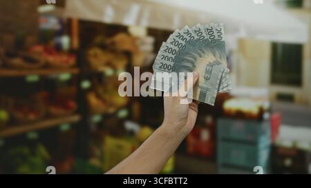 Kaukasischer Mann, der indonesische Rupiah-Banknoten in einem Supermarkt hält, mit Obst im Hintergrund als Symbol für Shopping und Geldwechsel. Stockfoto