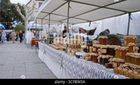 Frau, die auf einem lebhaften Straßenmarkt mit verschwommenem Hintergrund Ausschau hält und auf bunten Tischen unter weißen Zelten im geschäftigen U Gläser mit Honig und Konserven zeigt Stockfoto