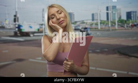 Frau drückt die Wange mit der Hand, die rosa Klemmbrett und silbernen Stift auf der belebten Straße hält; Unwohlsein. Stockfoto