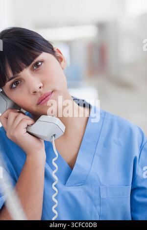 Krankenschwester, die im Krankenhausflur steht, trägt blaue Schürzen und hält Telefonhörer Stockfoto