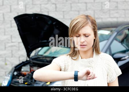 Frau, die Smartwatch überprüft, während sie neben einem kaputten Auto mit offener Motorhaube an einer Steinmauer steht Stockfoto