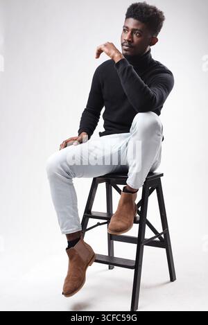 Lockiger, bärtiger, dunkelhäutiger Kerl im lässigen Outfit, der am Sessel sitzt und über den weißen Studiohintergrund blickt Stockfoto