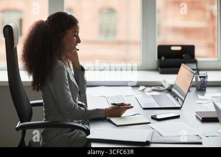 Junge Erwachsene Frau, die am Schreibtisch sitzt und im Büro arbeitet, Headset trägt, Stift hält, telefoniert, Computermonitor ansieht, Notizen auf Notizblock macht, von Dokumenten umgeben Stockfoto