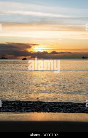 Ein atemberaubendes vertikales Bild fängt die Ruhe eines goldenen Sonnenaufgangs über dem Meer ein, mit einer Silhouette der Boote vor dem Horizont und den Wellen Stockfoto