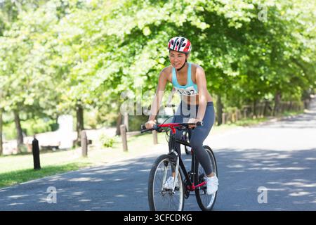 Asiatische Frau, die auf dem Parkweg mit schwarzem Fahrrad fährt, mit rot-weißem Helm und Lätzchen Nummer 452 Stockfoto