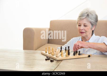 Ältere Frau, die sich auf Schachbrett konzentriert, während sie Schach auf Holztisch im Wohnzimmer spielt, Kopierraum Stockfoto