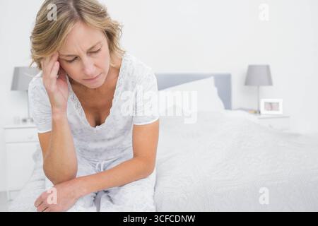 Ältere Frau mit Pyjama, die am Bettrand in der Nähe der Lampe im Schlafzimmer sitzt und den Tempel reibt Stockfoto