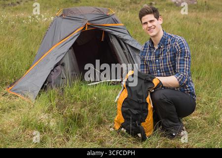 Mann kniet neben dem grauen, orangen Kuppelzelt auf einer grasbewachsenen Wiese, organisiert Rucksackausrüstung, Kopierraum Stockfoto