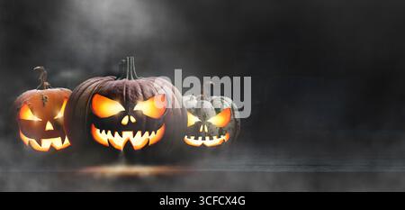 Eine Gruppe von drei gruseligen halloween Kürbislaternen, Jack o Laterne, mit leuchtenden Gesichtern auf einer hölzernen Plattform in einem dunklen gruseligen Kerker bei Nacht. Stockfoto