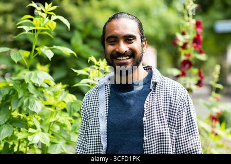 Mann lächelt in einem Garten mit üppigen grünen Pflanzen und hellen Blumen im Hintergrund. Deutschland Stockfoto