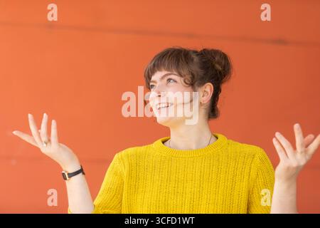 Frau in gelbem Pullover lächelt mit offenen Händen vor orangem Hintergrund. Deutschland Stockfoto