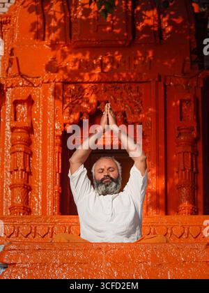 Ruhiger indischer Mann, der vor einem intensiven Orangentempel in Varanasi, Indien, meditiert und den inneren Frieden repräsentiert. Stockfoto