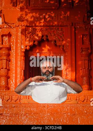 Ruhiger indischer Mann, der vor einem intensiven Orangentempel in Varanasi, Indien, meditiert und den inneren Frieden repräsentiert. Stockfoto