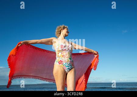 Reife Frau im Blumenbadeanzug, die roten Schal am Meer unter klarem blauem Himmel hält. Kepaniwai Park, Wailuku, Hawaii Stockfoto