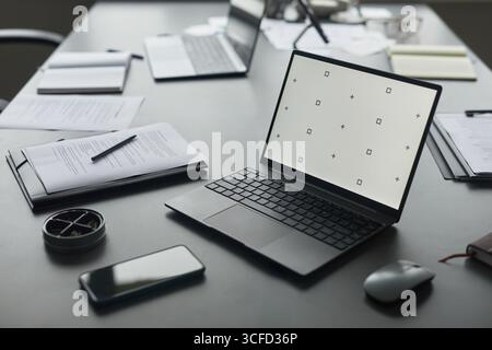 Moderner Bürotisch mit offenem Laptop mit leerem Bildschirm, Smartphone, Dokumenten, Notebooks und Stift, die über den gesamten Arbeitsbereich verteilt sind, was auf aktive geschäftliche oder externe Arbeitsumgebungen hindeutet Stockfoto