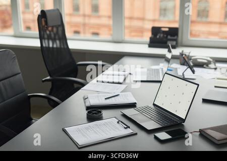 Moderner Büroarbeitsplatz mit offenem Laptop, Smartphone, Dokumenten, Stift und Kaffeetasse auf dem Schreibtisch in der Nähe eines großen Fensters, leeren ergonomischen Stühlen und zusätzlichen Laptops im Hintergrund Stockfoto
