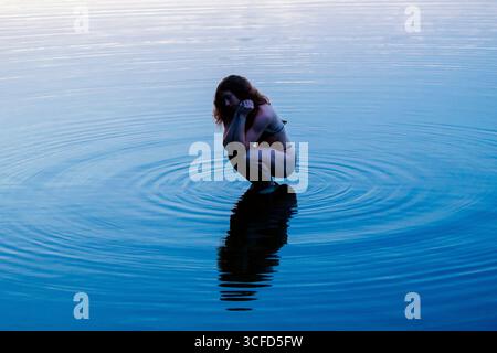 Frau, die sich in ruhigem blauem Wasser hockt und sanfte Wellen um ihr Spiegelbild kreiert. WA, USA Stockfoto