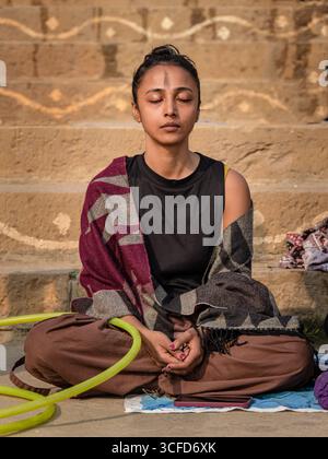 Junge Frau meditiert in Lotus-Position mit geschlossenen Augen auf den Ghats von Varanasi, Indien, die den inneren Frieden repräsentiert. Stockfoto