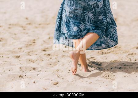 Nahaufnahme von Beinen und Füßen einer Frau, während sie barfuß auf weichem Sand läuft und ein blaues Sommerkleid trägt. Stockfoto