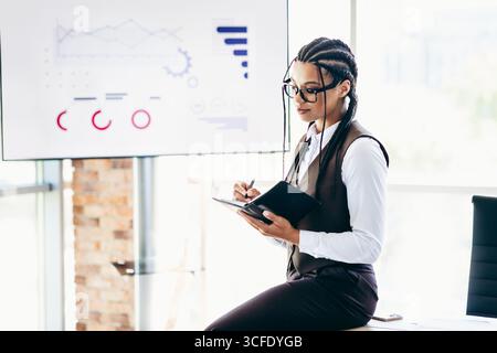 Professionelle Geschäftsfrau macht Notizen während einer strategischen Präsentation in einer modernen Büroumgebung mit Analysen Stockfoto