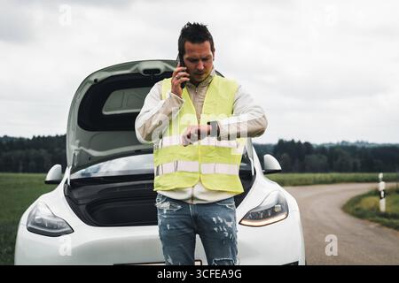 Bayern, Deutschland - 22. August 2025: Ein Mann in einer Warnweste spricht am Telefon vor seinem Pannenwagen auf einer Landstraße *** ein Mann mit Warnweste telefoniert vor seinem liegengebliebenen Auto auf einer Landstraße Stockfoto