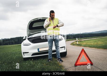 Bayern, Deutschland - 22. August 2025: Ein Mann in einer Warnweste spricht am Telefon vor seinem Pannenwagen auf einer Landstraße *** ein Mann mit Warnweste telefoniert vor seinem liegengebliebenen Auto auf einer Landstraße Stockfoto