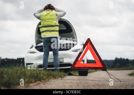Bayern, Deutschland - 22. August 2025: Symbolisches Bild einer Panne: Ein Mann steht verzweifelt mit geöffneter Motorhaube vor seinem Auto. Das Warndreieck ist aufgebaut *** Symbolbild Autopanne: Ein Mann steht verzweifelt vor seinem Auto mit geöffneter Motorhaube. Das Warndreieck ist aufgestellt Stockfoto