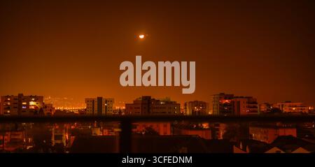 Mond sichtbar am orangefarbenen Nachthimmel über Wohngebäuden in Kiryat ATA, Israel, mit Stadtlichtern und städtischer Skyline Stockfoto