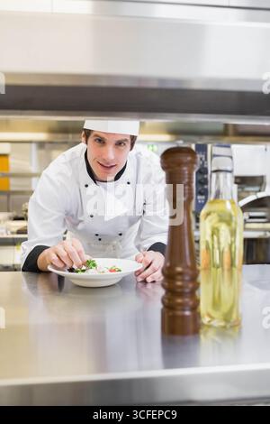 Männlicher Koch, der Salat auf weißem Teller in der kommerziellen Küche überzieht neben der Pfeffermühle Stockfoto