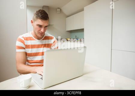 Mann, der am Küchentisch sitzt und sich auf silbernen Laptop mit Becher, Obstschale und Wasserflasche konzentriert Stockfoto